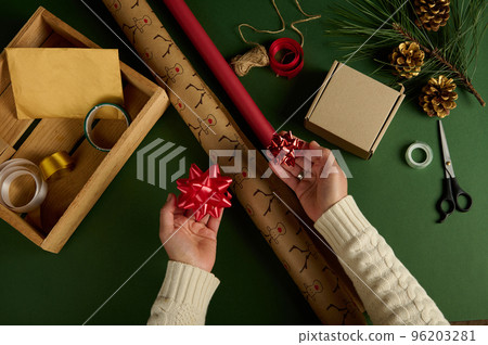 Top view woman's hands holding tied bows over wrapping paper with deer pattern and of red color, on a green surface with materials for packing Christmas and New Year gifts. Boxing Day. Handwork. Diy 96203281