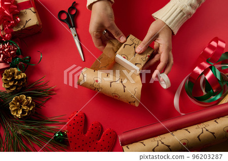 Diy presents. Xmas and New Year preparations. Step by step packing presents. Close-up of a craftswoman wrapping Christmas gift in a gift paper with deer pattern, tying a bow using white and red ribbon 96203287