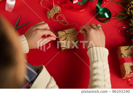 Close up. Overhead view woman in warm beige sweater, decorating Christmas presents, wrapped in deer pattern gift paper, making bow from linen rope . Boxing Day. Xmas and New Year celebration concept Close up. Overhead view woman in warm beige sweater, decorating Christmas presents, wrapped in deer pattern gift paper, making bow from linen rope . Boxing Day. Xmas and New Year celebration concept 96203290