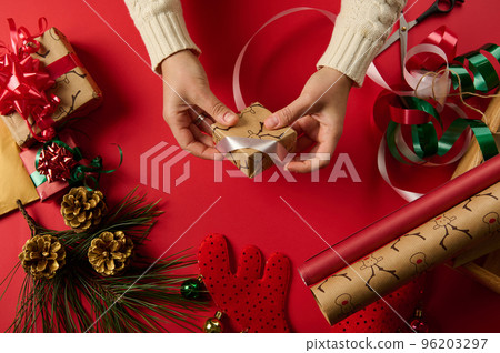 Diy presents. Xmas and New Year preparations. Step by step packing presents. Craftswoman wrapping Christmas gift in a gift paper with deer pattern, tying bow using white decorative ribbon. Close-up. 96203297