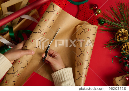 Top view of a woman in beige sweater, using scissors, cuts the wrapping gift paper with deer pattern over a red background, surrounded by wrapping materials and Christmas decorative items. Boxing Day Top view of a woman in beige sweater, using scissors, cuts the wrapping gift paper with deer pattern over a red background, surrounded by wrapping materials and Christmas decorative items. Boxing Day 96203318