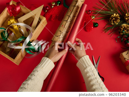 Top view of female hands picking up wrapping paper and decorative ribbon, while packing Christmas or New Year gift on red surface. Xmas preparations. Boxing day. Happy winter holidays. Celebration 96203340