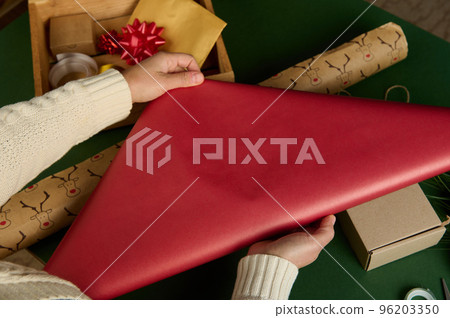 Top view woman's hands rolling out red gift paper over green surface with laid out wrapping decorative materials, bows, tapes and ribbons. Diy presents, Handwork art. New Years, Christmas preparations Top view woman's hands rolling out red gift paper over green surface with laid out wrapping decorative materials, bows, tapes and ribbons. Diy presents, Handwork art. New Years, Christmas preparations 96203350