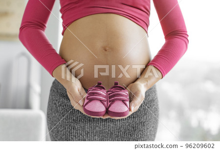 Pregnancy. Pregnant woman expecting baby girl holding pink red shoes in gender reveal. Expecting mom showing baby bump belly. Baby clothing Pregnancy. Pregnant woman expecting baby girl holding pink red shoes in gender reveal. Expecting mom showing baby bump belly. Baby clothing 96209602