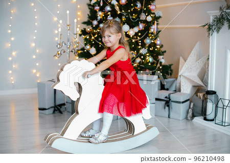 Christmas,x-mas,winter concept - girl in red dress sitting on wood horse toy in New Year's decorations 96210498