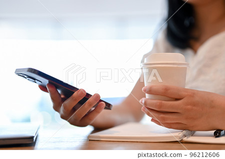 Asian woman using her smartphone and sipping coffee while relaxing in the coffee shop. cropped Asian woman using her smartphone and sipping coffee while relaxing in the coffee shop. cropped 96212606