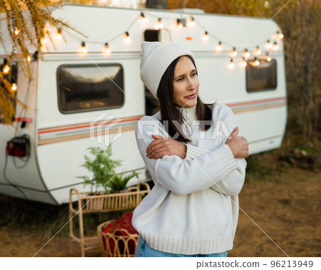 A pensive Caucasian woman in a white knitted sweater and a hat is resting near a motorhome in a warm autumn. Travel by camper. A pensive Caucasian woman in a white knitted sweater and a hat is resting near a motorhome in a warm autumn. Travel by camper. 96213949