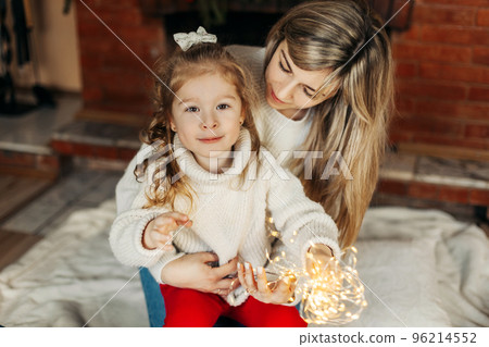 Portrait of a happy mother and daughter with Christmas lights on the background of the fireplace. The concept of Christmas and New Year. Time together, Christmas time Portrait of a happy mother and daughter with Christmas lights on the background of the fireplace. The concept of Christmas and New Year. Time together, Christmas time 96214552