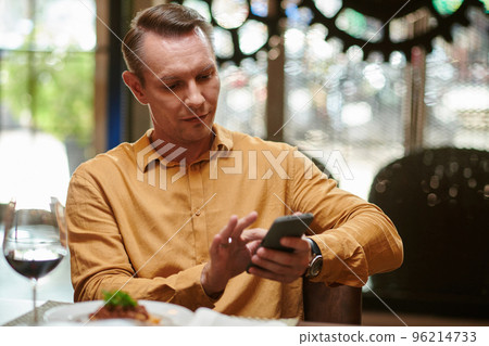 Smiling man sitting at table in restaurant and checking social media on smartphone 96214733