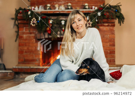 A smiling young woman is playing with a dog, sitting on the floor near the fireplace, on Christmas Eve. Christmas mood, Christmas holidays. The concept of Christmas and New Year 96214972