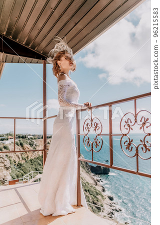 Romantic bride, blond girl in white wedding dress with open shoulders posing on open terrace with backdrop of the sea and rocks. Stylish young woman standing on terrace and looking on ocean at sunset Romantic bride, blond girl in white wedding dress with open shoulders posing on open terrace with backdrop of the sea and rocks. Stylish young woman standing on terrace and looking on ocean at sunset 96215583