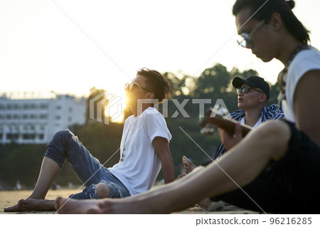 three young asian adult men sitting on sand beach playing guitar at dusk 96216285