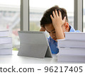Young Asian elementary school boy sitting next to stack of books and using tablet computer with both hands holding his head with gestures and face showing stress. 96217405