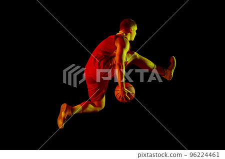 Studio shot of young active athlete, male basketball player in sports uniform in motion and action with ball isolated over dark background in neon light 96224461