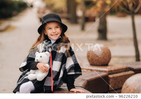 A little stylish girl in autumn clothes is sitting on a bench in the autumn city 96226210