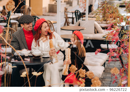 A stylish family of three is sitting at a table outside in a cafe and drinking coffee. Dad, mom and daughter in the autumn city 96226722