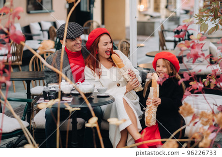 A stylish family of three is sitting at a table outside in a cafe and drinking coffee. Dad, mom and daughter in the autumn city 96226733
