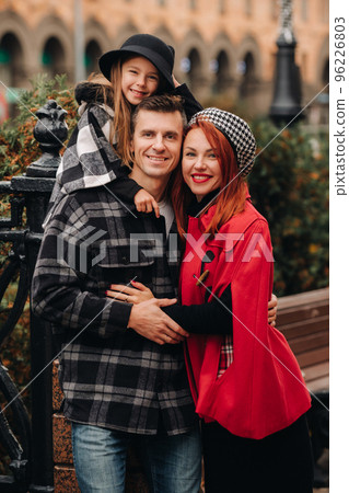 A stylish family of three strolls through the autumn city posing for a photographer . Dad, mom and daughter in the autumn city A stylish family of three strolls through the autumn city posing for a photographer . Dad, mom and daughter in the autumn city 96226803
