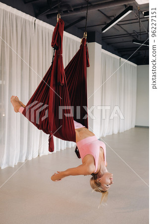 A young girl, an aero yoga coach, is hanging on a hammock in the yoga club hall. a woman in pink sportswear is doing anti-gravity yoga 96227151