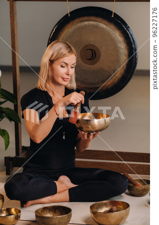A woman plays a Tibetan singing bowl while sitting on a yoga mat against the background of a gong 96227176