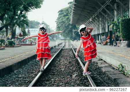 Two little sisters in red dresses walk along the train tracks in summer. Sisters were walking on the tracks at the train station. Travel and transportation concepts. 96227267