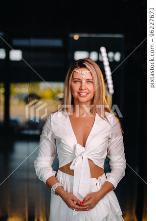 A positive young woman in a white dress and an ornament on her head smiles at the camera while standing in the studio 96227671