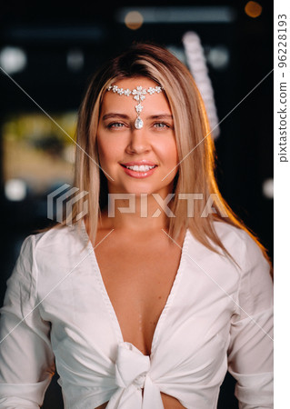 A positive young woman in a white dress and an ornament on her head smiles at the camera while standing in the studio 96228193