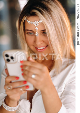 A positive young woman in a white dress and an ornament on her head was smiling, holding a phone in her hands and looking into it 96228274