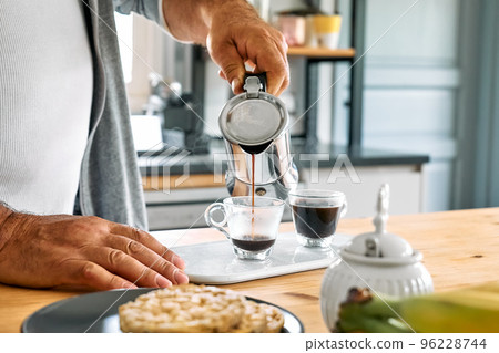 Man preparing classic Italian coffee in the mocha in the kitchen. Pouring coffee from moka pot into small glass coffee cup. Coffee brake. Morning habit. Man preparing classic Italian coffee in the mocha in the kitchen. Pouring coffee from moka pot into small glass coffee cup. Coffee brake. Morning habit. 96228744