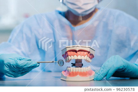 A dental doctor wearing blue gloves and a mask holds a dental model of the upper and lower jaws and a dental mirror 96230578