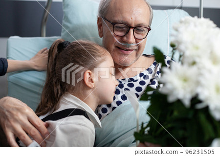 Girl hugging old man, lying in hospital bed. Senior, granddaughter showing physical affection each other in geriatric clinic observation room. Happy elderly patient receiving surprise from child. 96231505