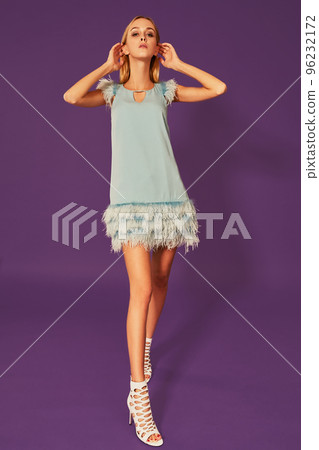 Image of cute beautiful slim blonde woman in elegant cocktail blue dress is posing at studio on purple background. Image of cute beautiful slim blonde woman in elegant cocktail blue dress is posing at studio on purple background. 96232172