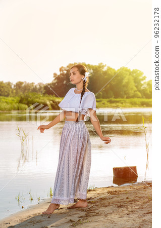 On the edge of the shore near the river stands a girl in a white dress. On the edge of the shore near the river stands a girl in a white dress. 96232178