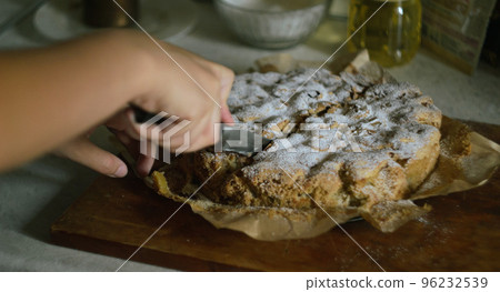 A woman's hand cuts a ready-made Charlotte apple pie into equal parts with a knife in the kitchen on a wooden board 96232539
