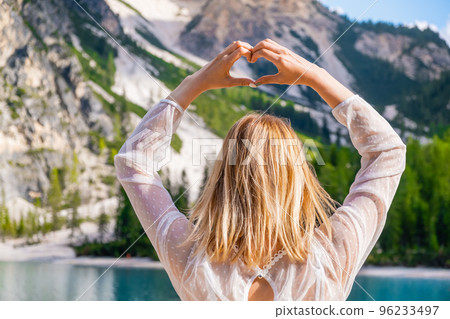 Blonde hair woman shows the heart with her hands on the background of the Dolomites and Lake Braies. 96233497