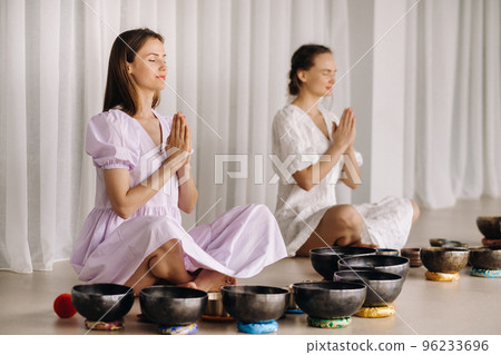 Two women are sitting with Tibetan bowls in the lotus position before a yoga class in the gym 96233696