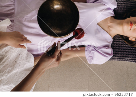 The copper singing bowl of the Nepalese Buddha in the spa. A young beautiful woman is doing a massage with singing bowls in a spa salon against the backdrop of a waterfall 96233717
