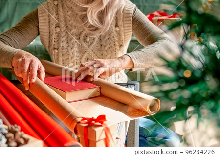 Wrapping christmas gifts. Blond woman wrapping presents in recycled card and decorated it with dried oranges and fir branches near the Christmas tree. Winter holiday celebration. 96234226