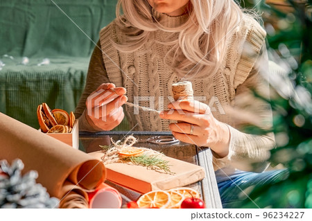 Wrapping christmas gifts. Blond woman wrapping presents in recycled card and decorated it with dried oranges and fir branches near the Christmas tree. Winter holiday celebration. Wrapping christmas gifts. Blond woman wrapping presents in recycled card and decorated it with dried oranges and fir branches near the Christmas tree. Winter holiday celebration. 96234227