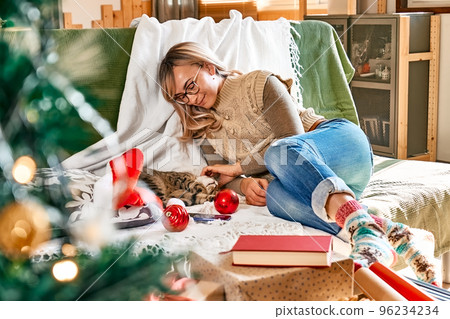 Cheerful blonde woman in festive mood hugging sleeping tabby cat on couch while preparing for celebration Christmas or New Year. Winter holiday concept. Cheerful blonde woman in festive mood hugging sleeping tabby cat on couch while preparing for celebration Christmas or New Year. Winter holiday concept. 96234234
