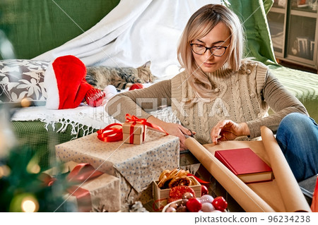 Wrapping christmas gifts. Blond woman wrapping a book as present in recycled card and decorate it with dried oranges and fir branches near the Christmas tree. Winter holiday celebration. 96234238