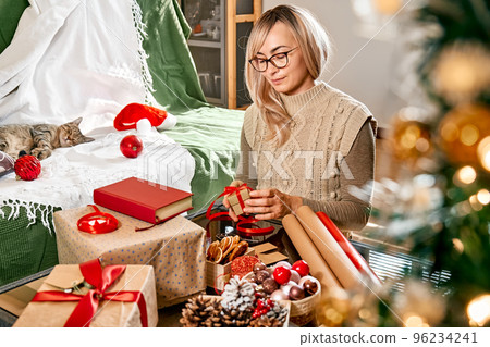 Wrapping christmas gifts. Blond woman wrapping present in recycled card and decorate it with red ribbon, dried oranges and fir branches near the Christmas tree. Winter holiday celebration. Wrapping christmas gifts. Blond woman wrapping present in recycled card and decorate it with red ribbon, dried oranges and fir branches near the Christmas tree. Winter holiday celebration. 96234241
