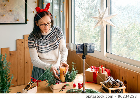 Female florist blogger making winter ikebana with fresh pine branches, candle and christmas decorations in streaming in front of her smartphone. Small business. Seasonal winter workshop. Female florist blogger making winter ikebana with fresh pine branches, candle and christmas decorations in streaming in front of her smartphone. Small business. Seasonal winter workshop. 96234258
