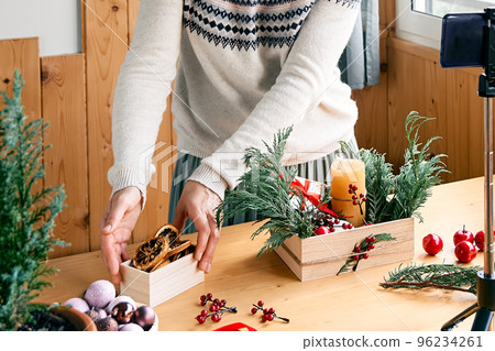 Female florist blogger making winter ikebana with fresh pine branches, candle and christmas decorations in streaming in front of her smartphone. Small business. Seasonal winter workshop. 96234261