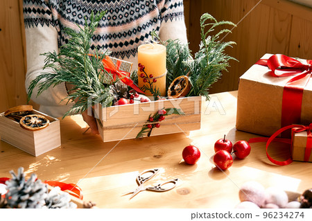 Woman's hands making christmas arrangement. Female florist creating winter ikebana with fresh pine branches, candle and christmas decorations. Small business. Seasonal winter workshop. 96234274
