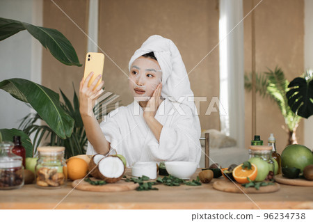 Close up portrait of happy asian woman blogger holding phone applying white cotton mask sheet 96234738