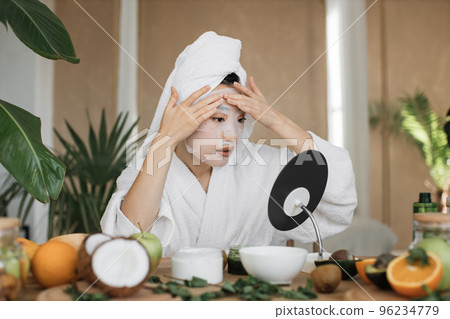 Attractive asian woman looking at mirror sitting at table with ingredients for homemade cosmetics Attractive asian woman looking at mirror sitting at table with ingredients for homemade cosmetics 96234779