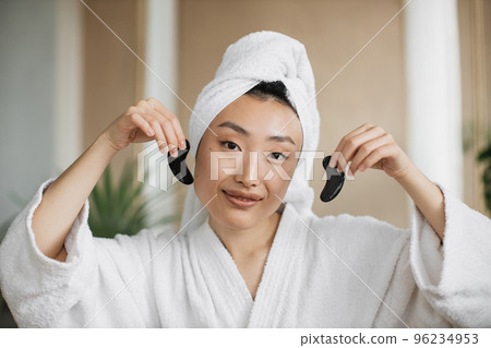 Pleasant asian woman in white bathrobe and towel holding eye patches Pleasant asian woman in white bathrobe and towel holding eye patches 96234953