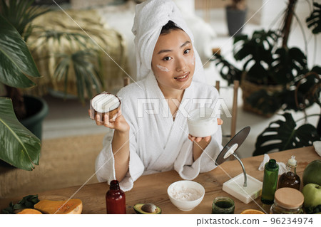 Young asian woman in white bathrobe and towel holding half of coconut and making homemade cream 96234974