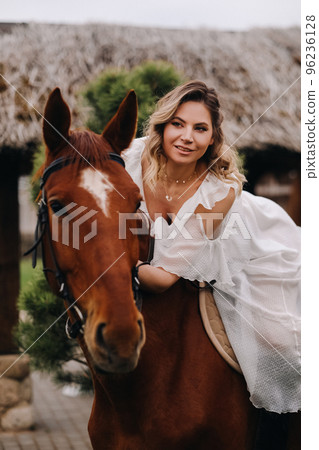 A woman in a white sundress riding a horse near a farm 96236128
